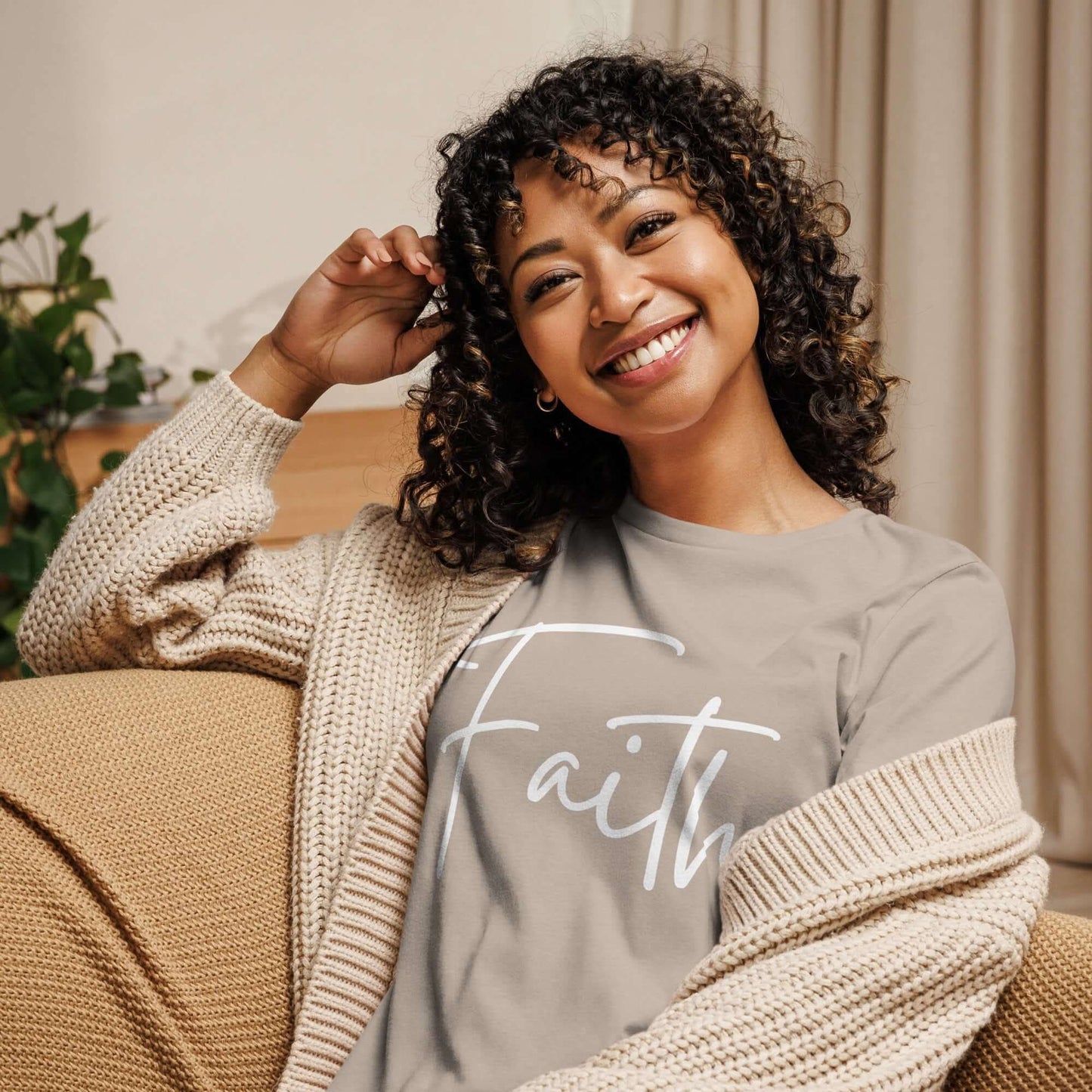 Woman smiling while wearing the Faith tee in white cursive lettering, cozy in a cardigan at home.