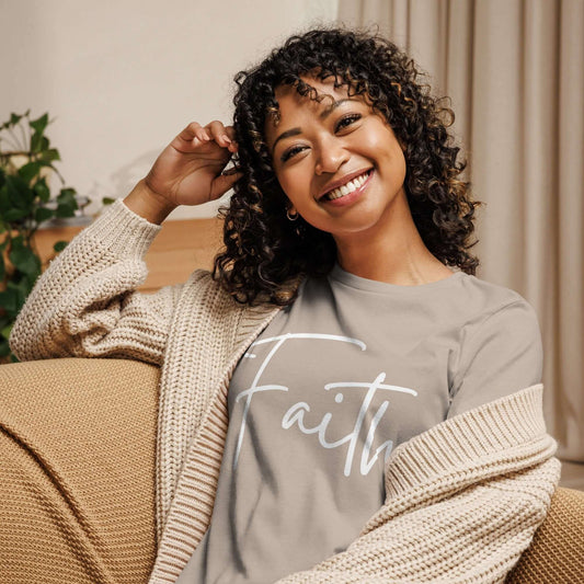 Woman smiling while wearing the Faith tee in white cursive lettering, cozy in a cardigan at home.