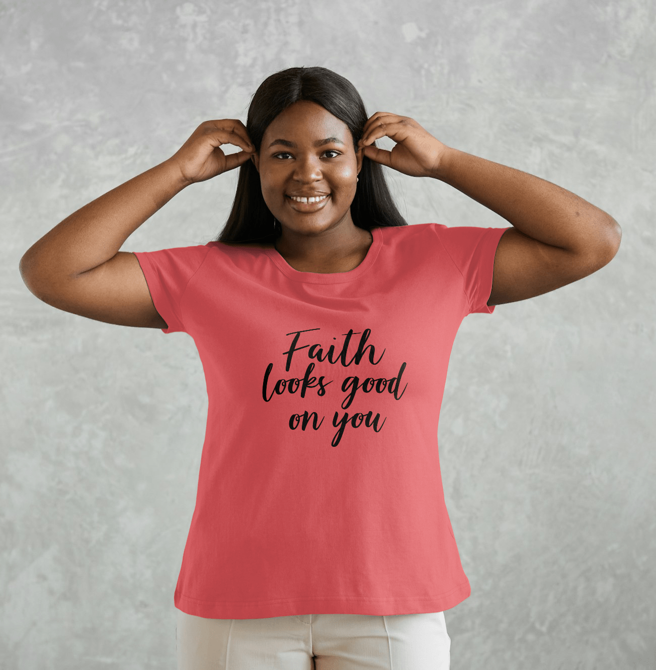 Woman wearing a pink tee with the phrase 'Faith Looks Good on You Tee' in cursive lettering, smiling.